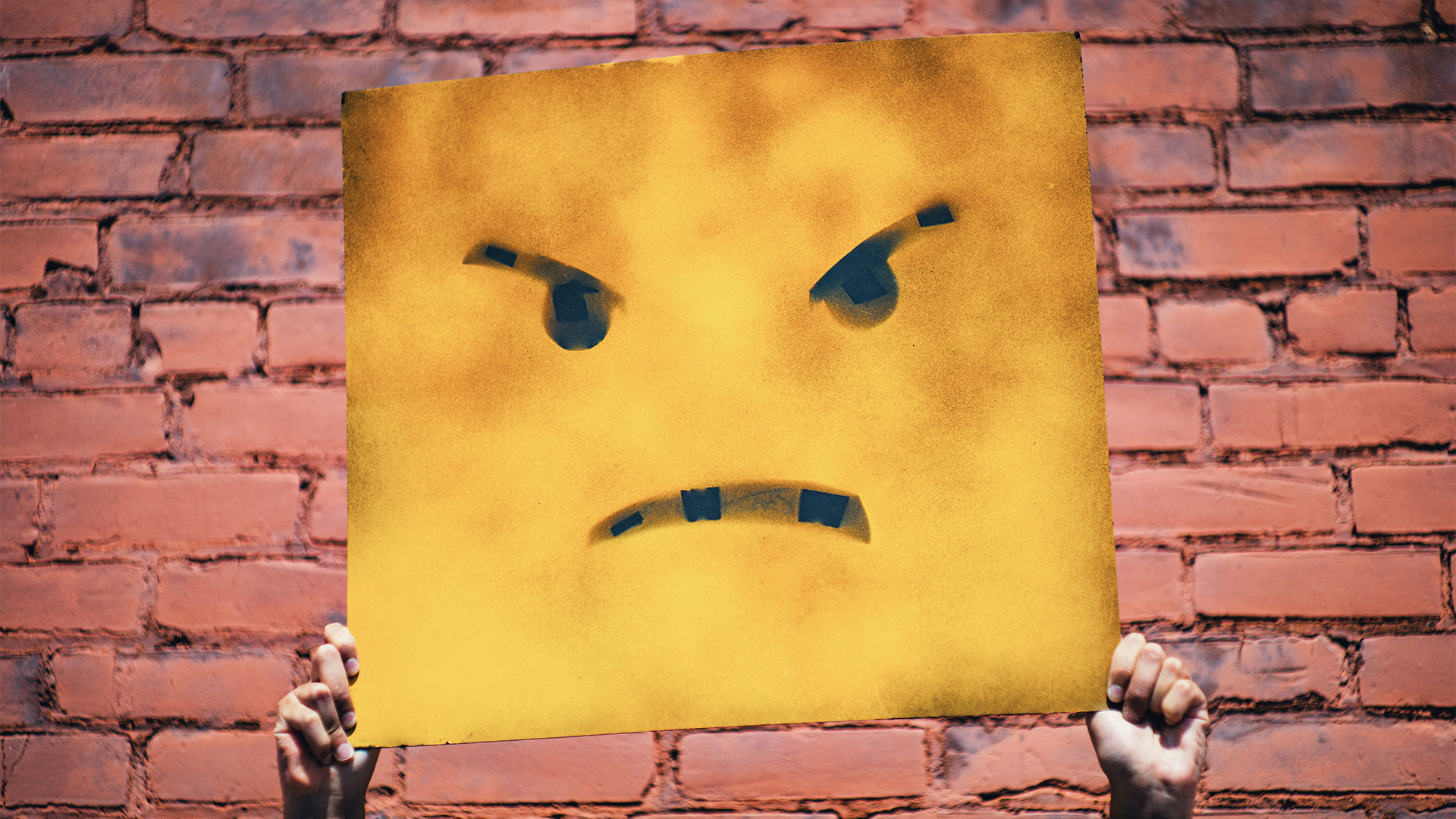 En person håller upp en tavla eller skylt med en arg emoji på